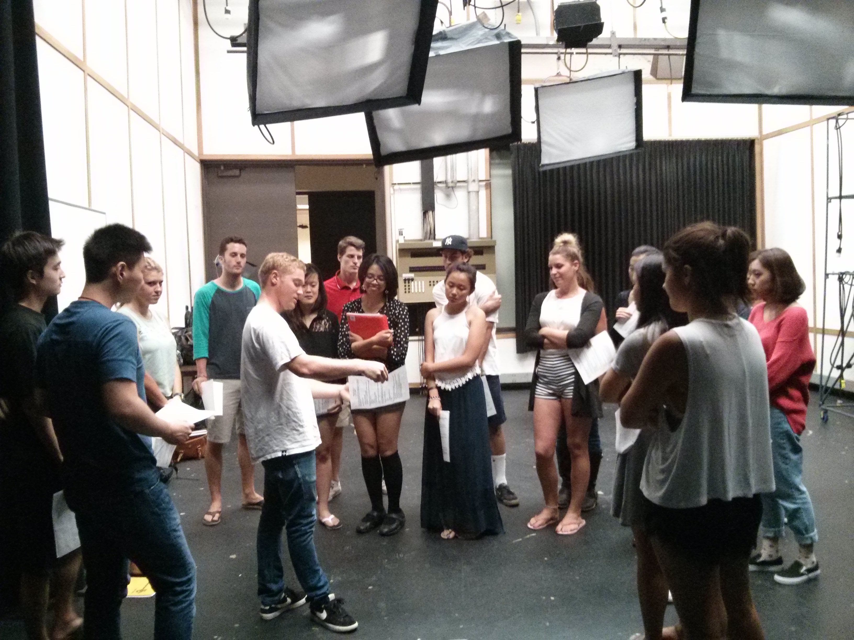 a dozen college college students stand in a circle discussing plans for filming in a studio