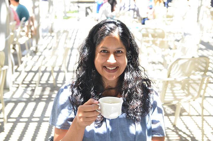 Gayatri sits under dappled light with a cup of coffee smiling